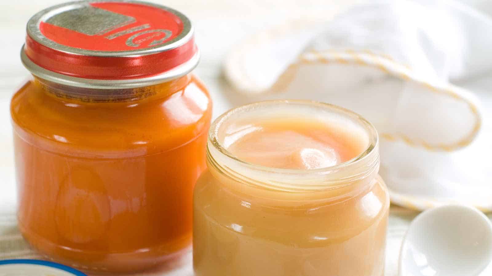 Two glass jars of baby food, one with orange puree and a closed red lid, the other with light beige puree and an open lid, placed on a white surface.