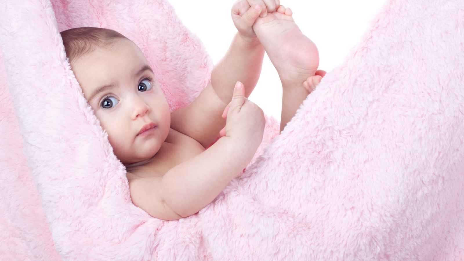 A baby lies in a pink, fluffy blanket, holding one foot with both hands and looking toward the camera.