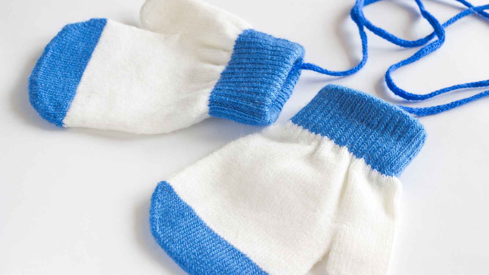 A pair of white knitted mittens with blue cuffs and tips, connected by a blue string, lying on a white surface.