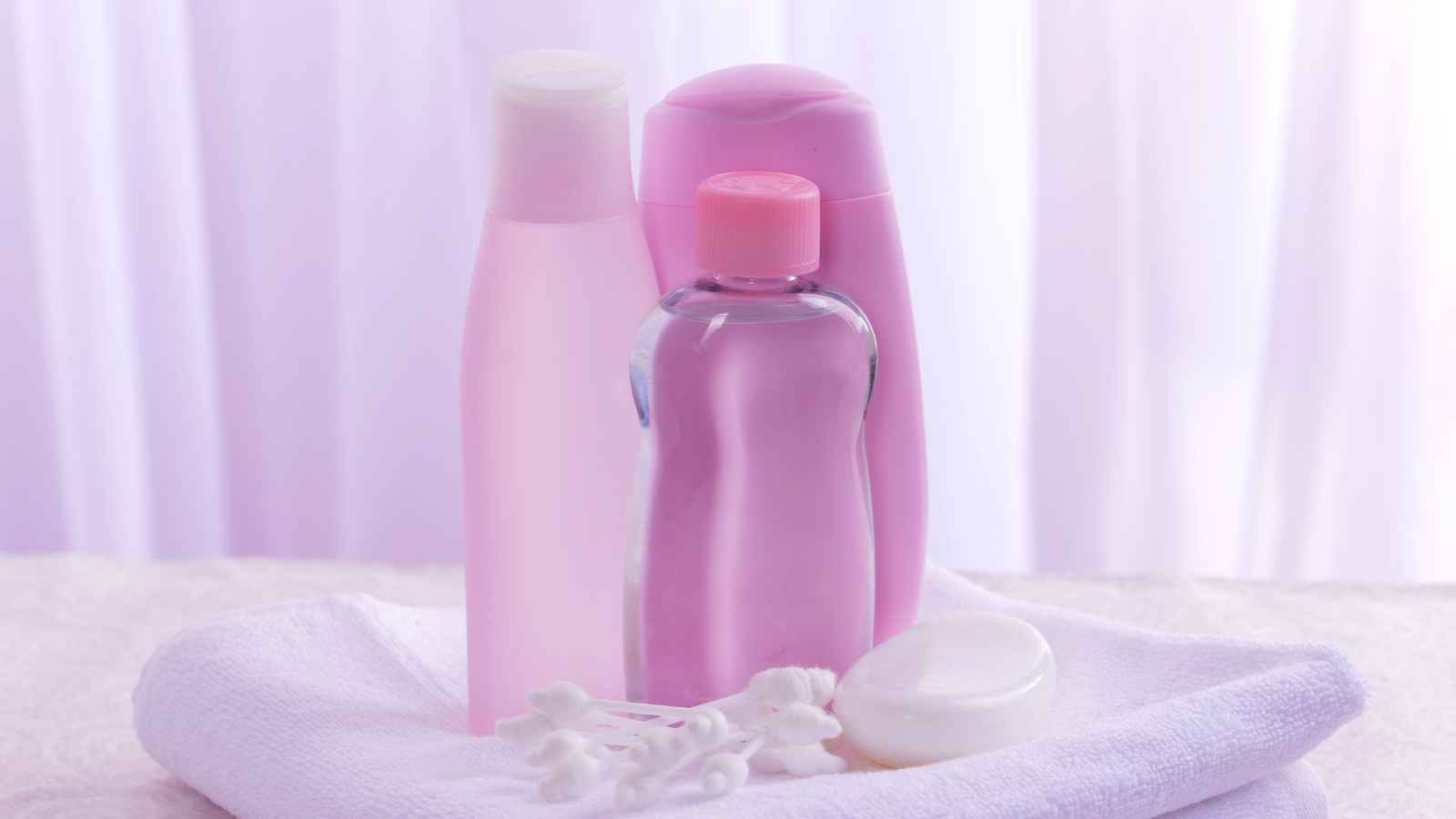 Three pink and clear toiletry bottles, a white round soap, and cotton swabs are arranged on a white towel with a soft, light background.