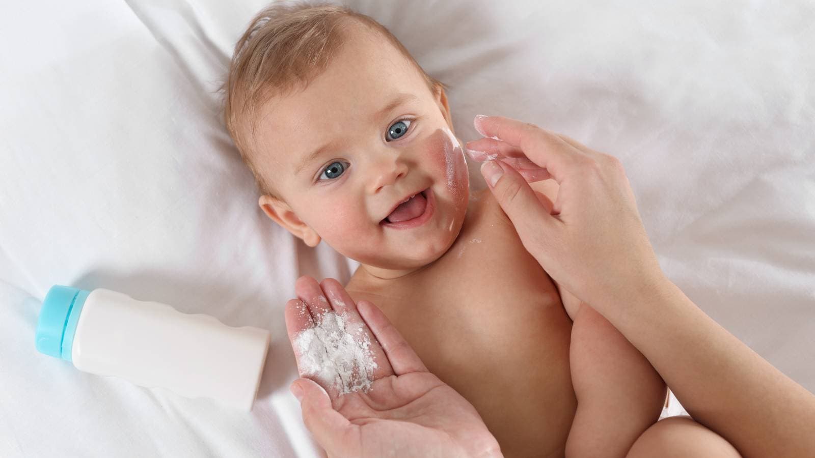 A baby lying on a white surface is smiling while an adult applies baby powder to its cheek. A bottle of baby powder is visible nearby.