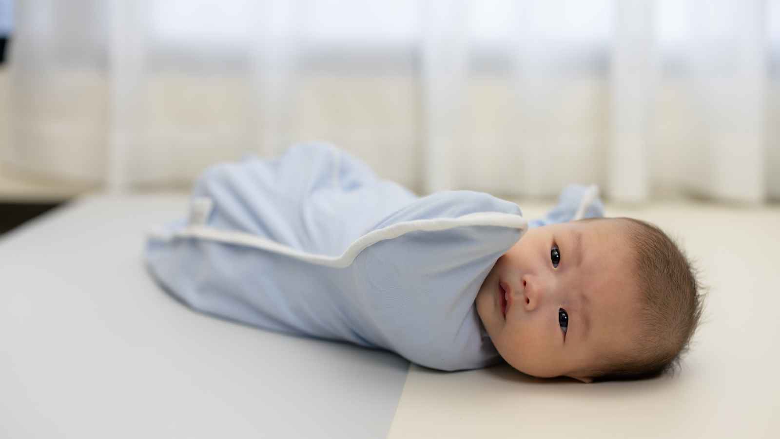 A baby wrapped in a light blue blanket lies on a surface, looking towards the camera with a neutral expression. White curtains are in the background.