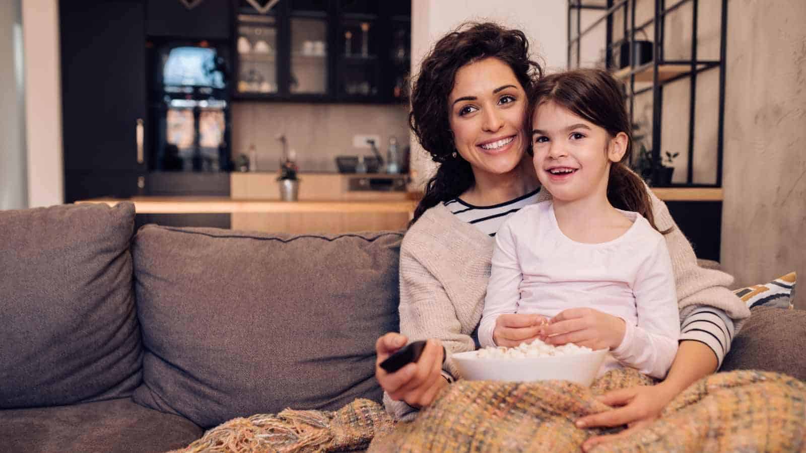 A woman and a young girl sit on a couch under a blanket, smiling and holding a bowl of popcorn, with a remote control and a kitchen in the background.