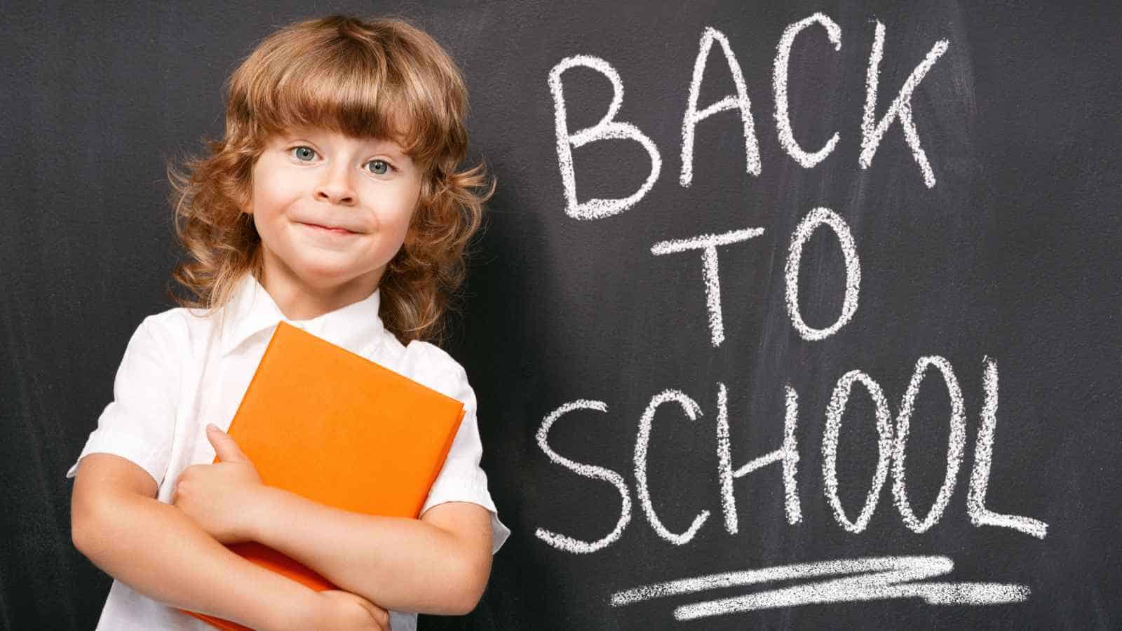 Young child holding an orange book stands in front of a blackboard with "Back to School" written in large white letters.