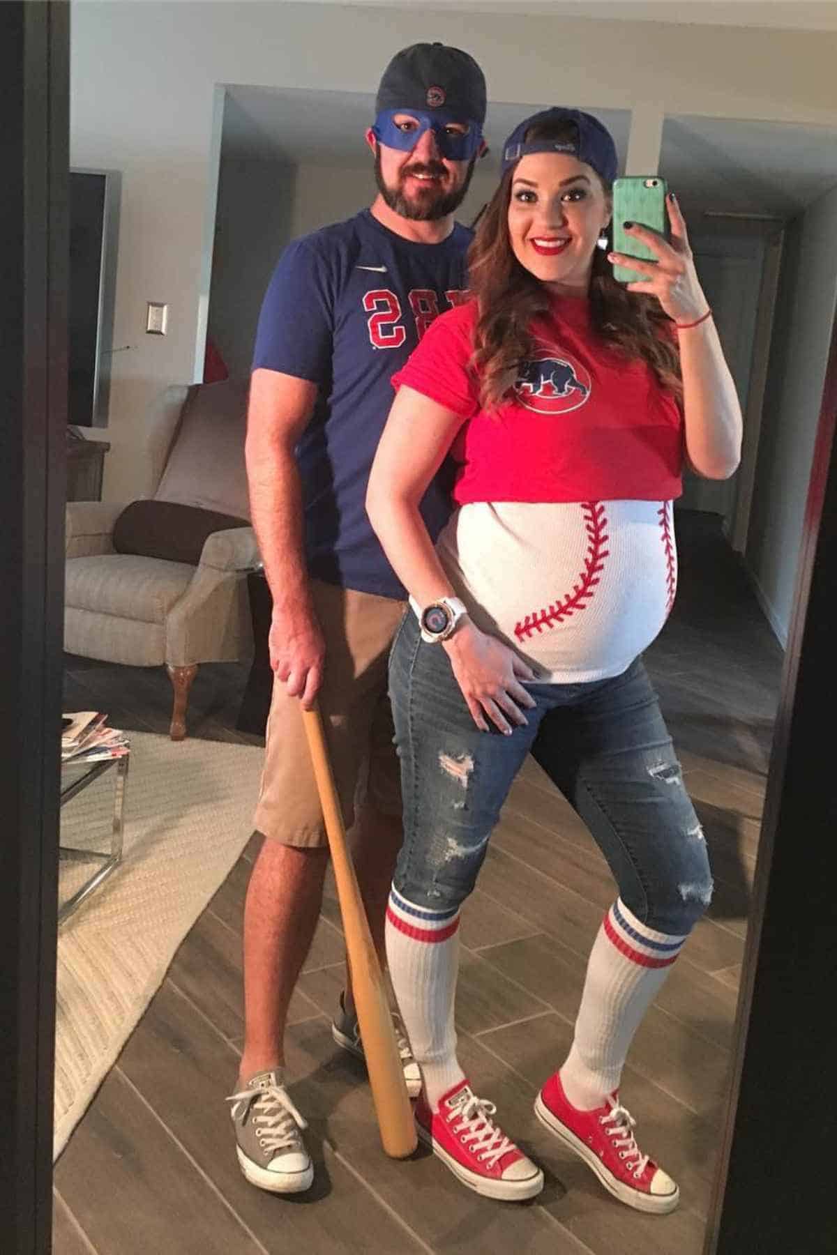 A couple takes a mirror selfie; the man is dressed as a baseball player and the woman, who is pregnant, is wearing a shirt and painted belly resembling a baseball.