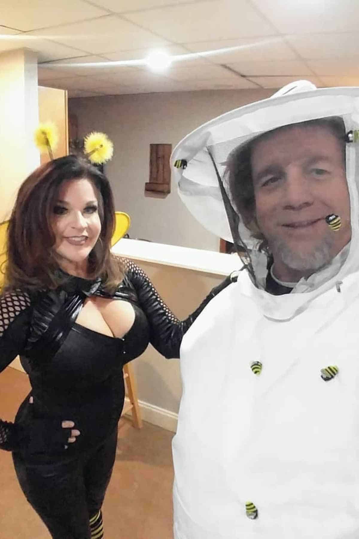 A woman in a bee costume stands next to a man dressed as a beekeeper, both smiling indoors with bee decorations on the beekeeper suit.