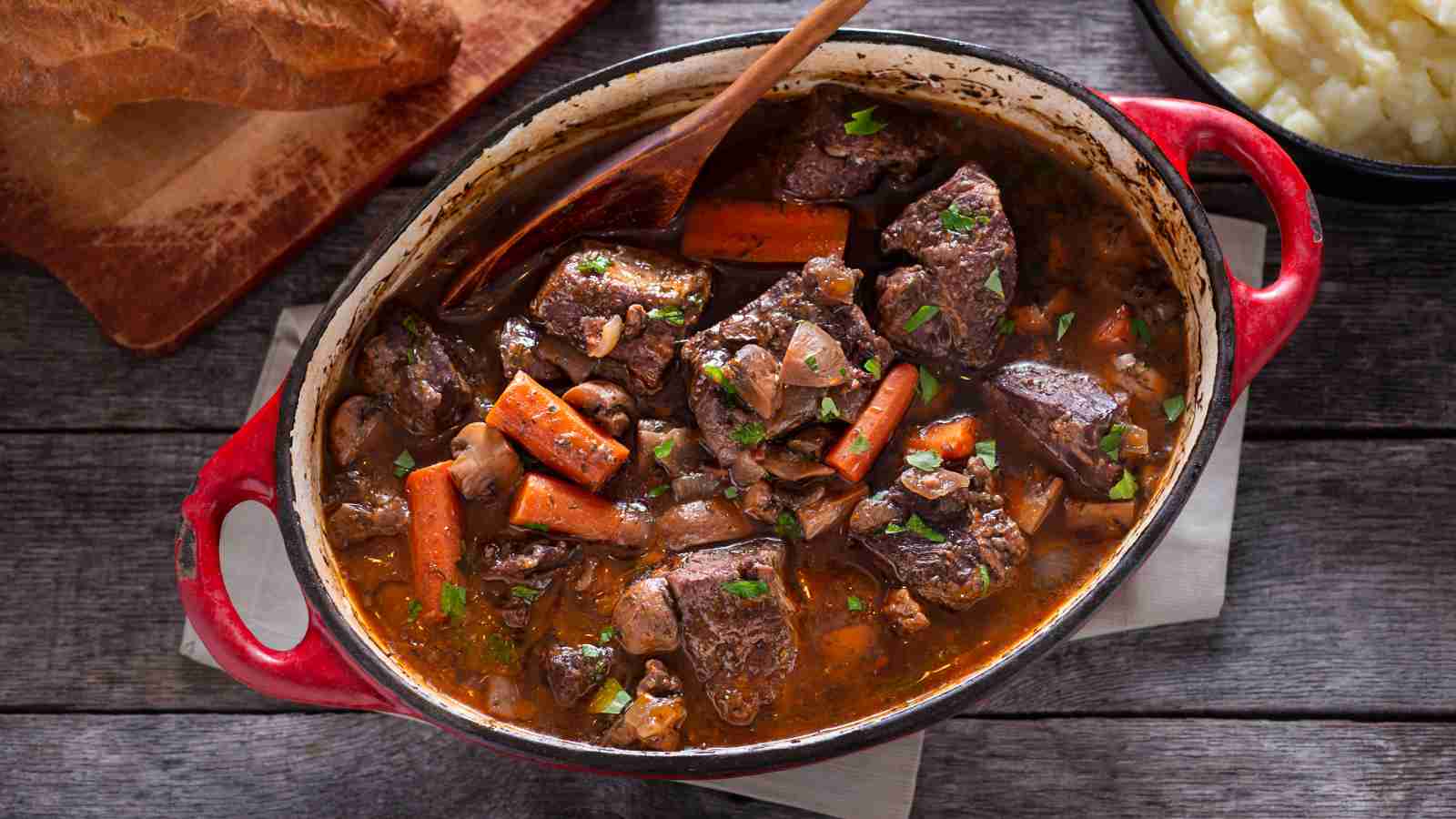 A red pot filled with beef stew, featuring chunks of beef, carrots, mushrooms, and herbs, sits on a wooden table beside bread and mashed potatoes.