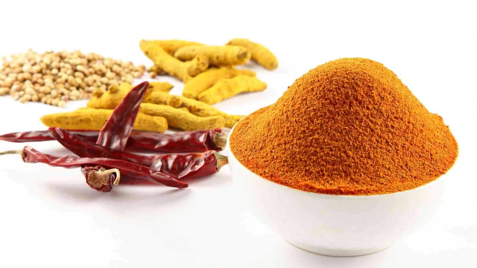 A white bowl filled with berbere is placed next to dried red chilies, turmeric roots, and coriander seeds on a white background.