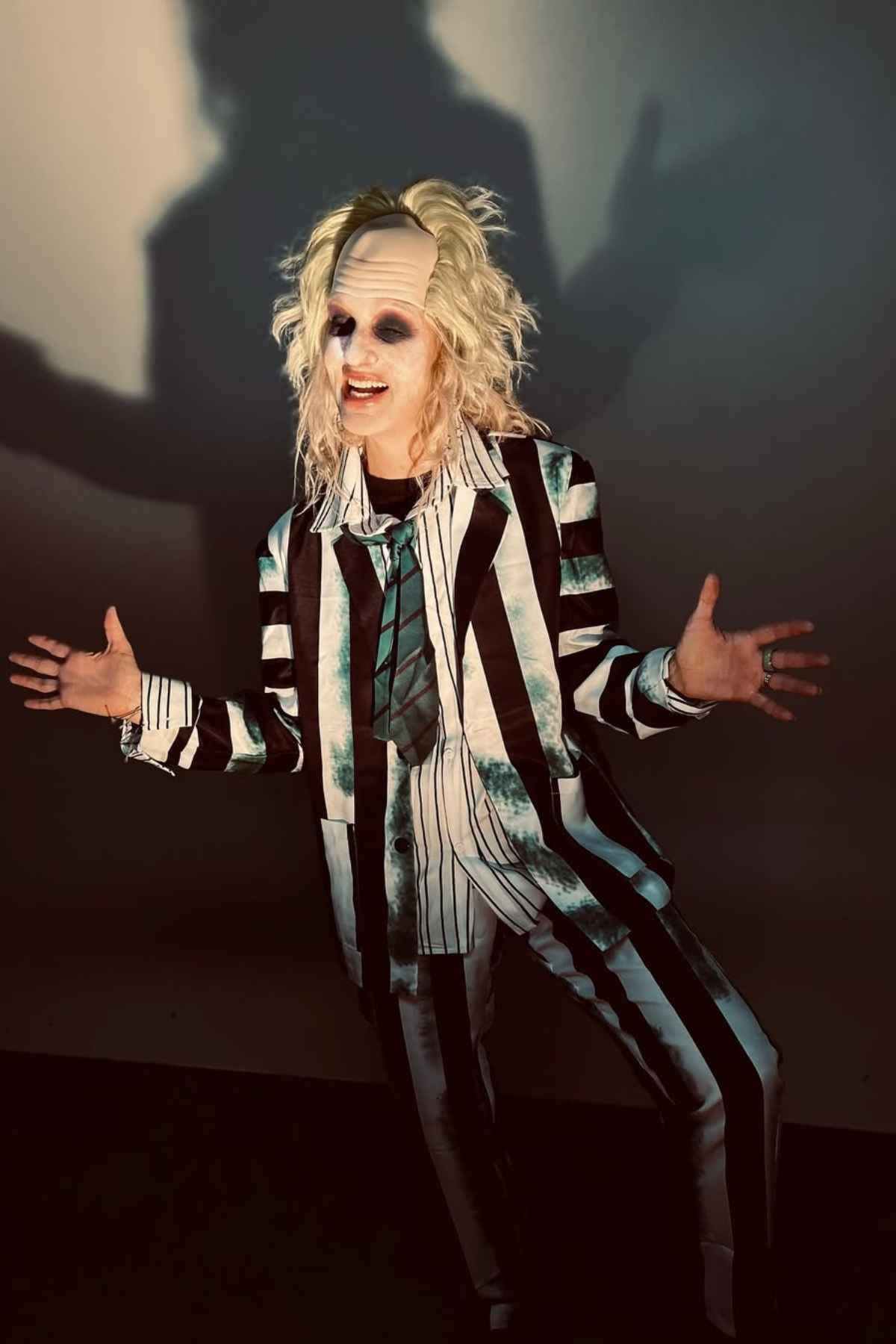 A person dressed as Beetlejuice, wearing a black and white striped suit with disheveled blond hair, pale makeup, and dark eye circles, standing in front of a shadowed wall.