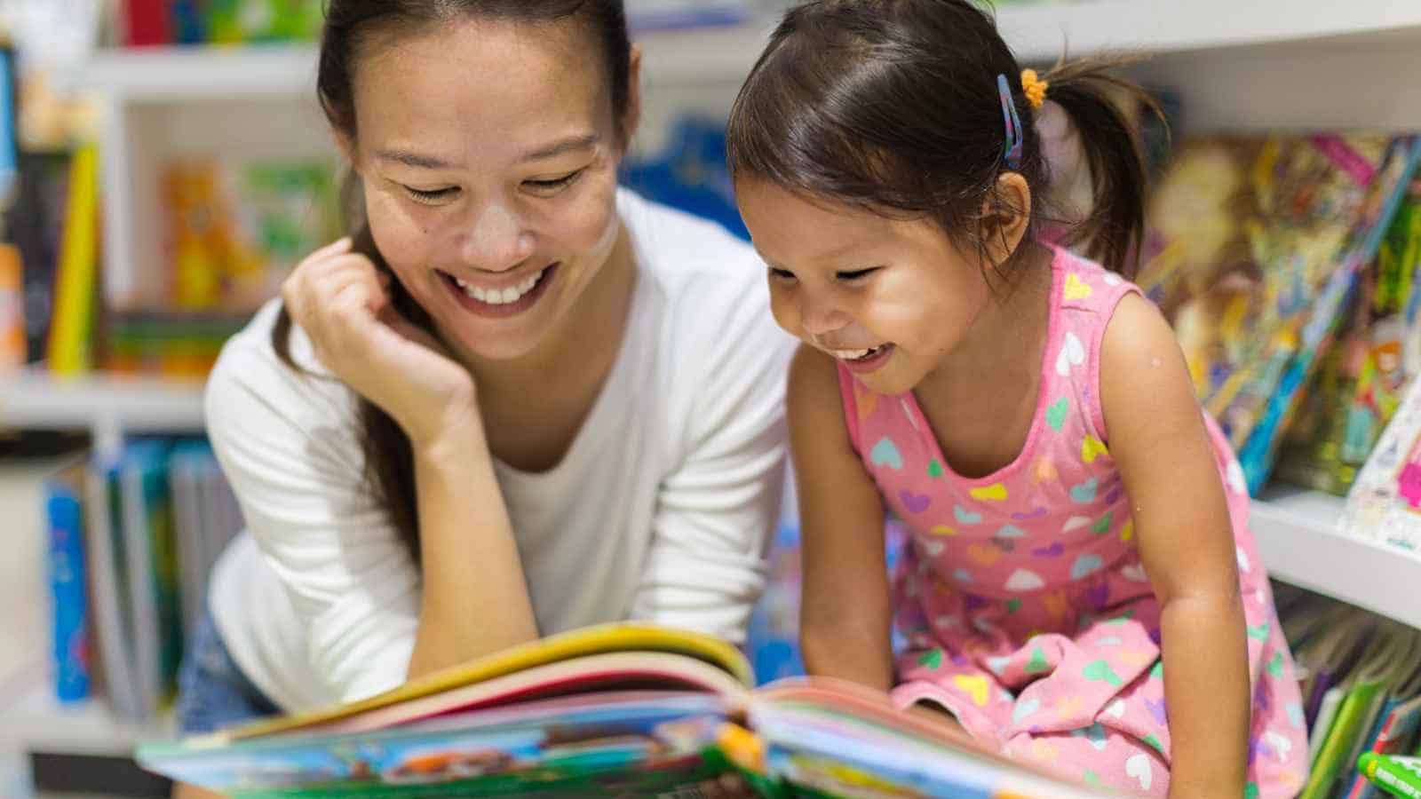 An adult and a child sit together in a library or bookstore, smiling and reading a colorful book. Shelves filled with books are visible in the background.