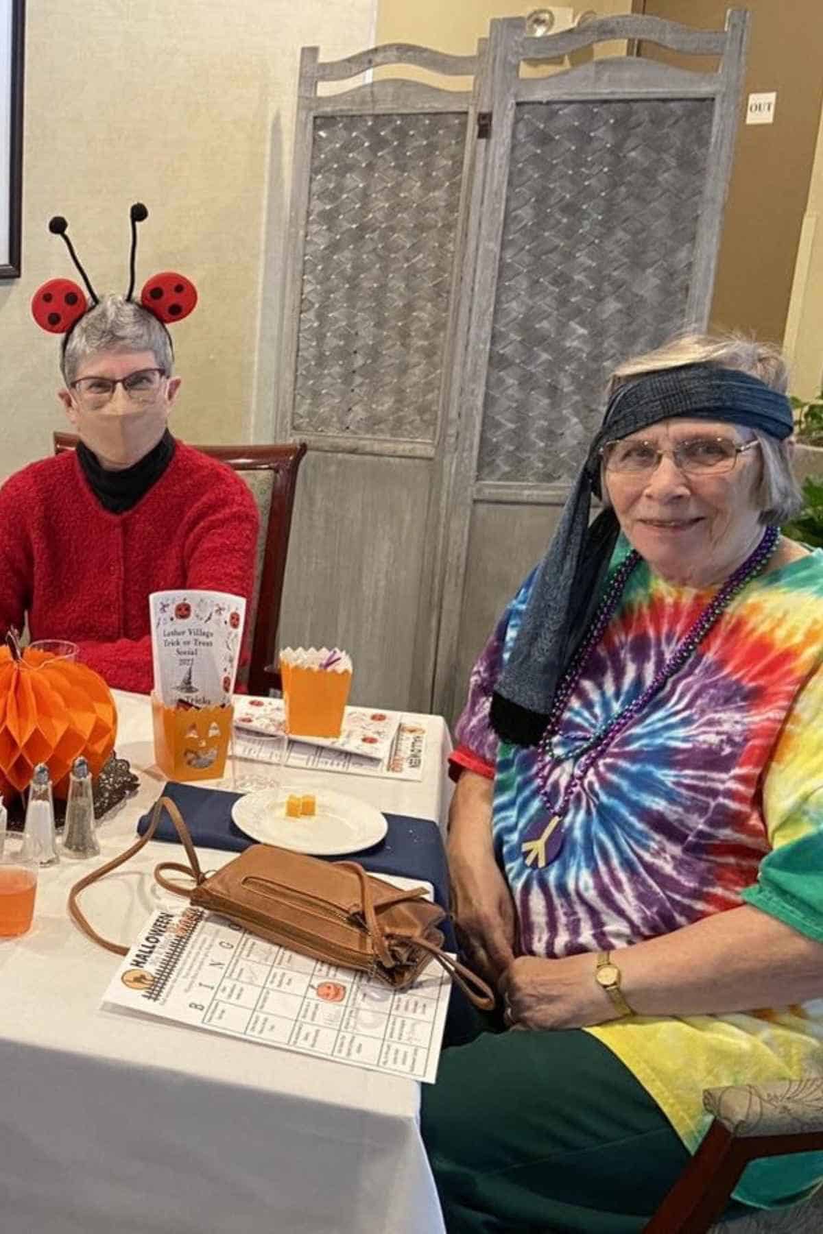 Two elderly sit at a table; one wears ladybug antennae and a red sweater, the other a tie-dye shirt and headband. Halloween decorations and bingo cards are on the table.