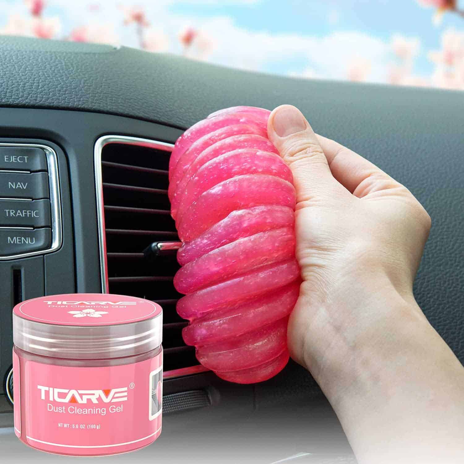 A hand uses pink cleaning gel to clean the air vent of a car. A container labeled "TICARVE Dust Cleaning Gel" is placed in the foreground.