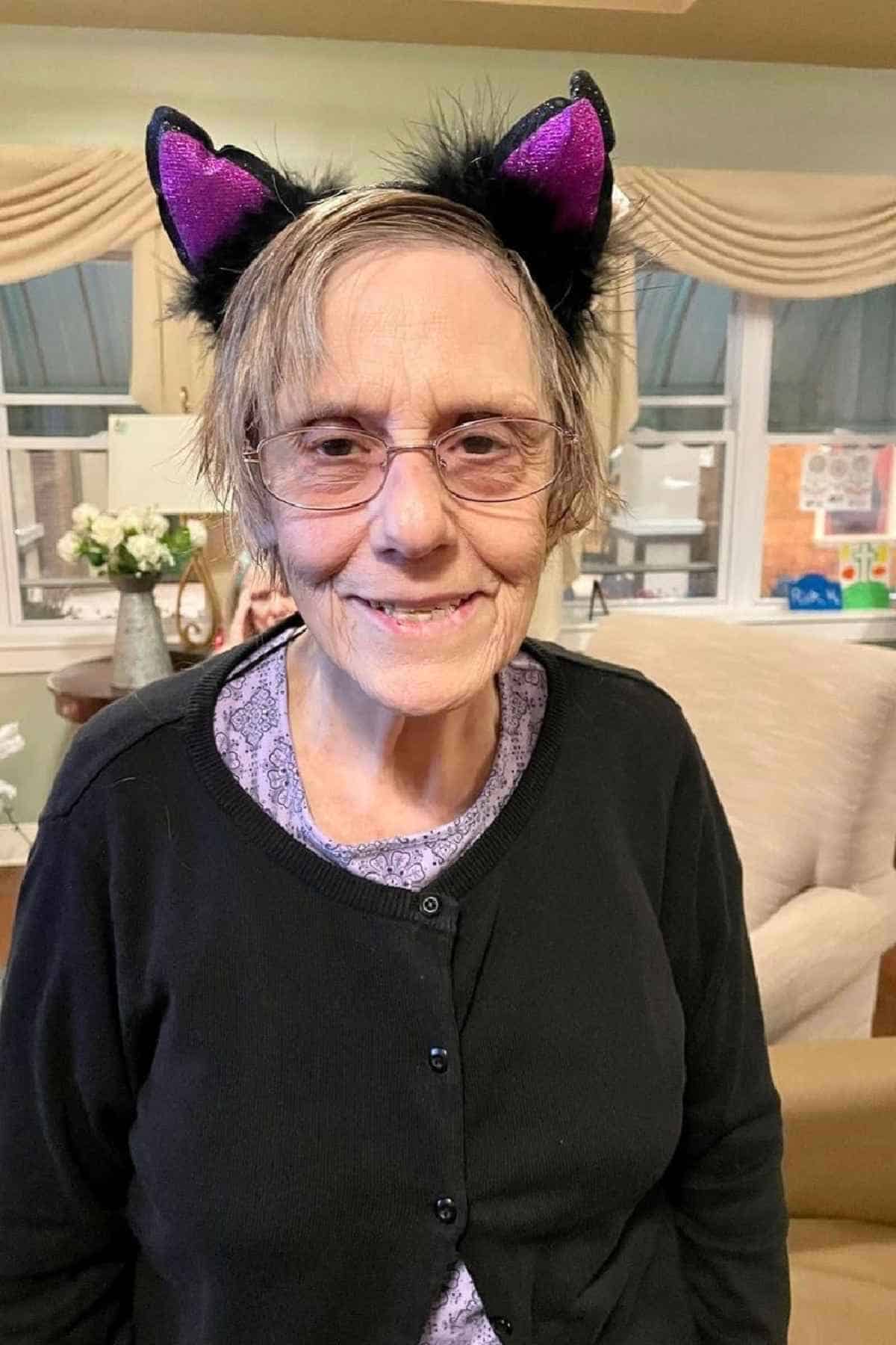 An older woman wearing glasses, a black cardigan, and a headband with black and purple cat ears, standing indoors and smiling at the camera.