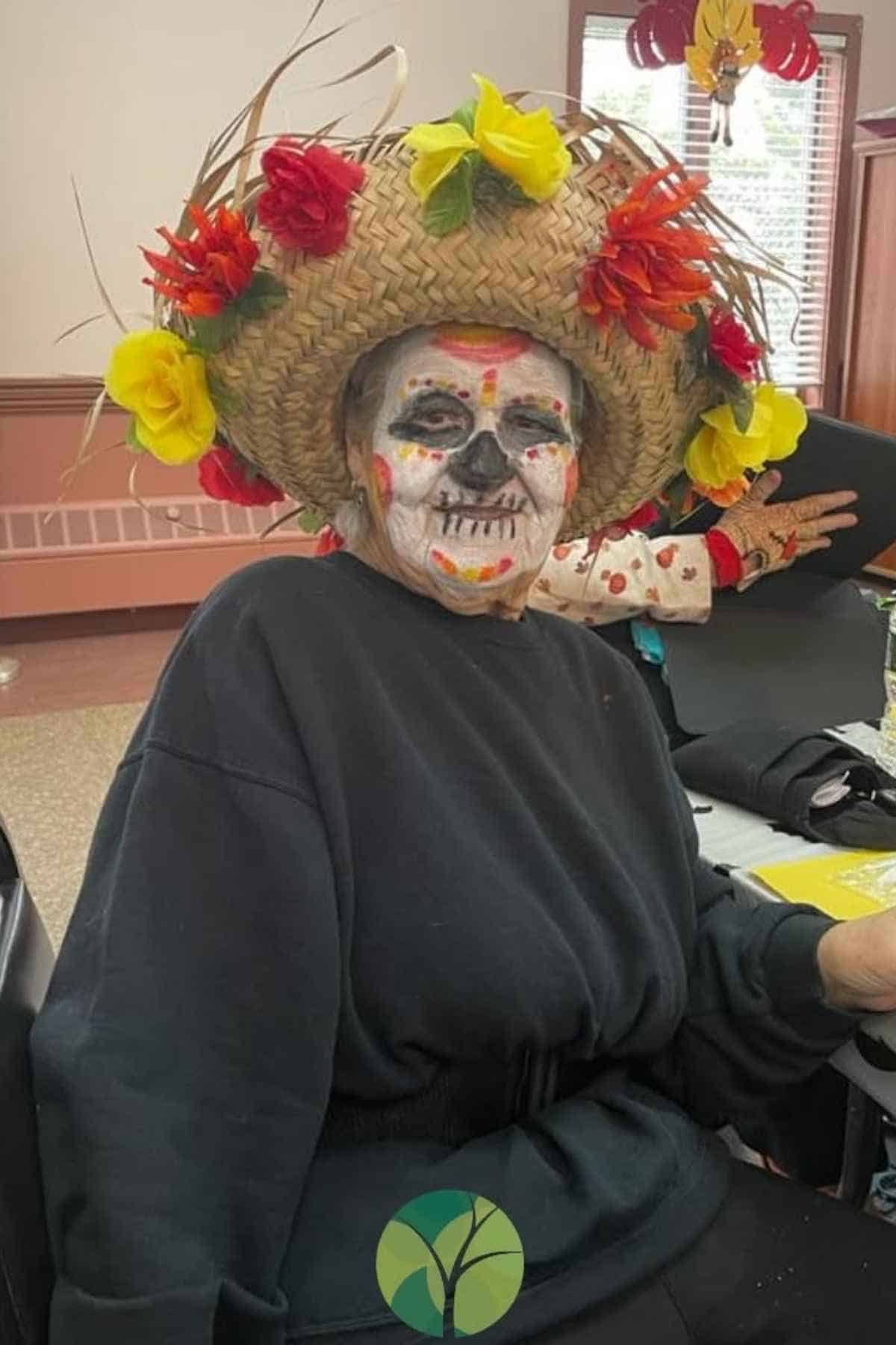 Person wearing a large straw hat decorated with colorful flowers and face painted in a skull design, sitting indoors.
