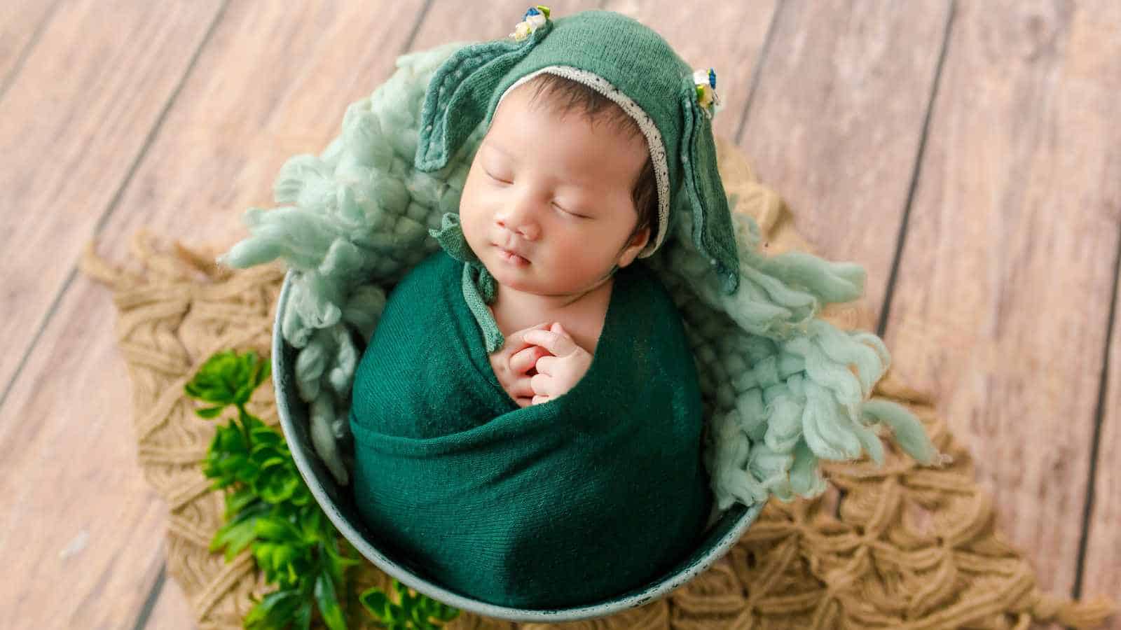 A sleeping baby wrapped in a green blanket and hat lies in a bowl on a wooden floor, surrounded by textured fabric and greenery.