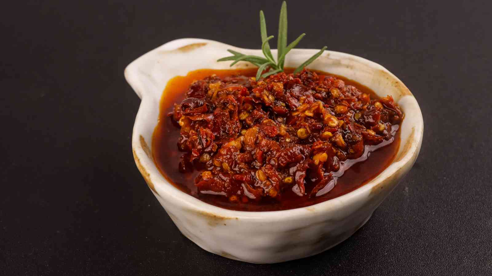 A small white bowl filled with chili crisp garnished with a sprig of rosemary, set against a black background.