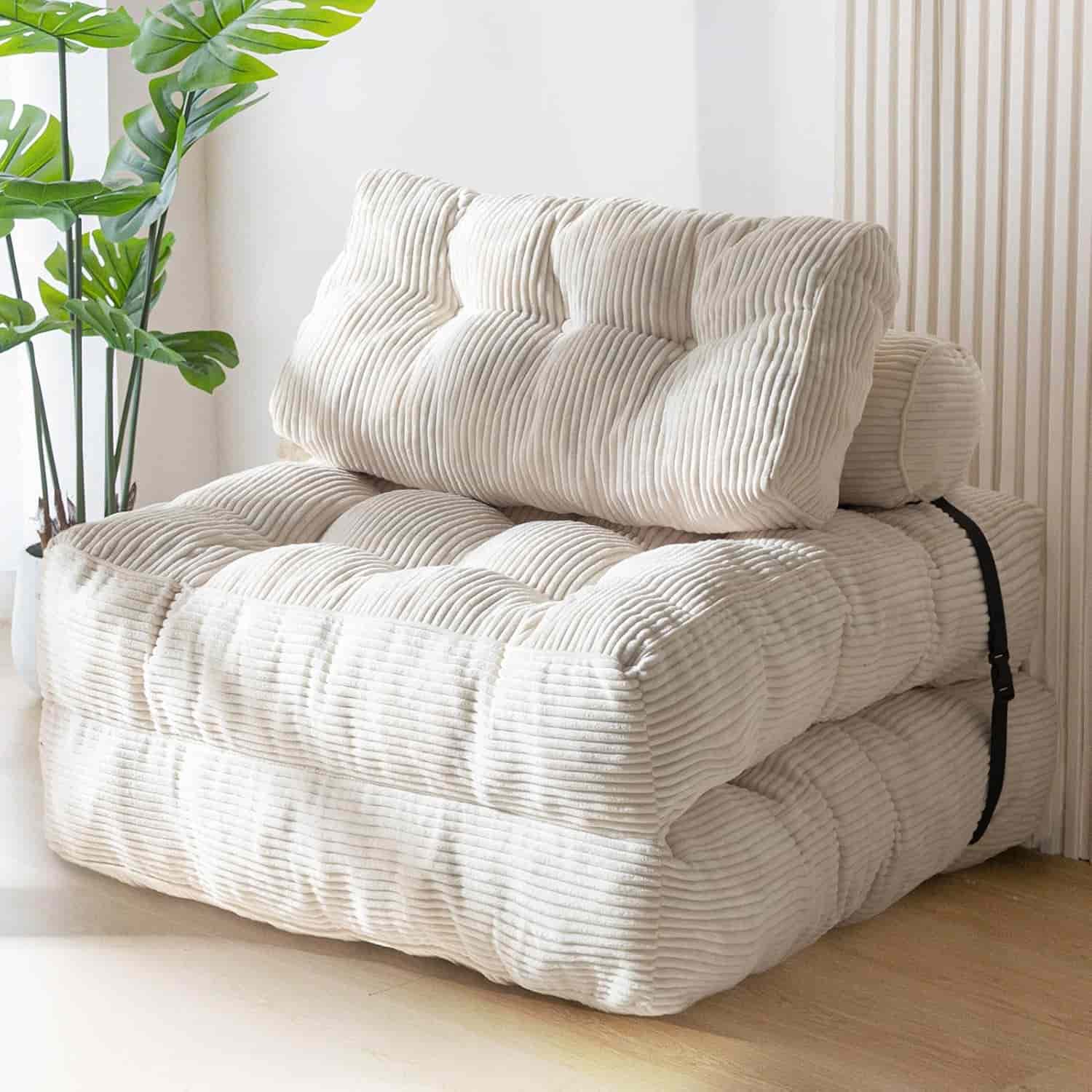 A cream-colored, tufted corduroy floor chair with a matching back cushion is folded and strapped together, placed beside a green potted plant in a bright room.