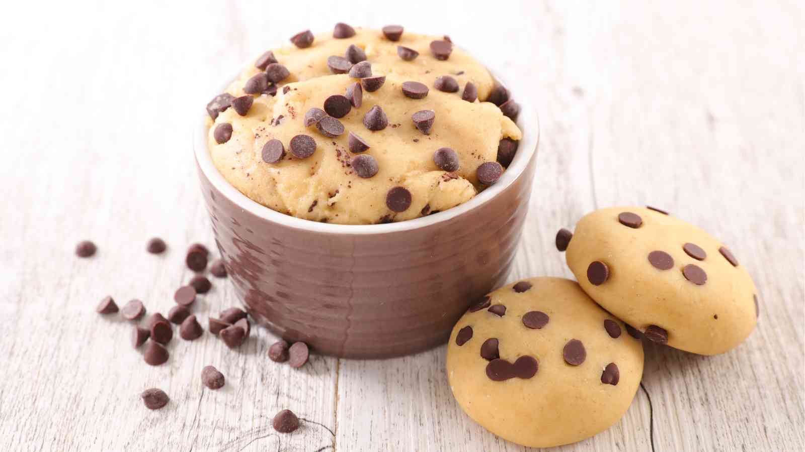 A brown ramekin filled with chocolate chip cookie dough and topped with chocolate chips sits next to two chocolate chip cookies and scattered chips on a light wooden surface.