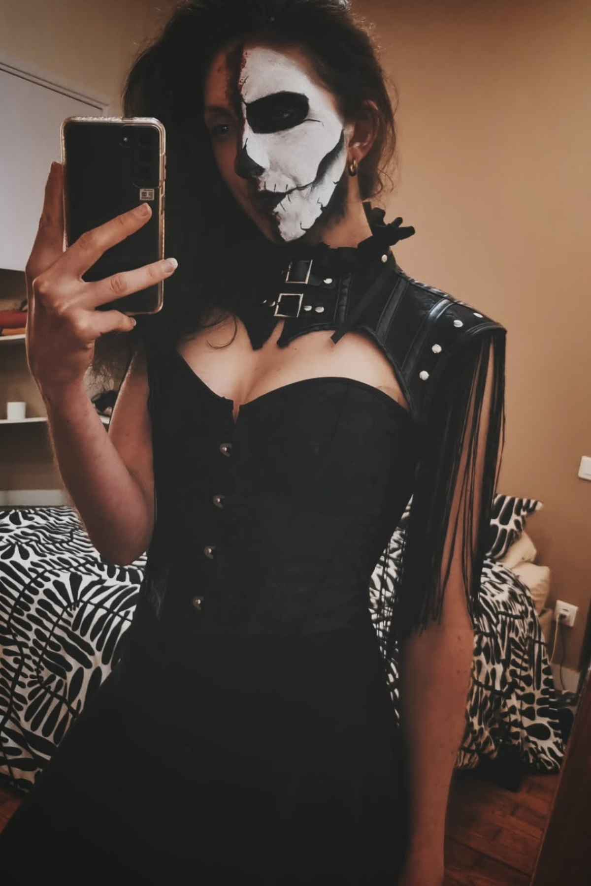 Person taking a mirror selfie wearing a black corset outfit, with one half of their face painted white and black in a skeletal design.
