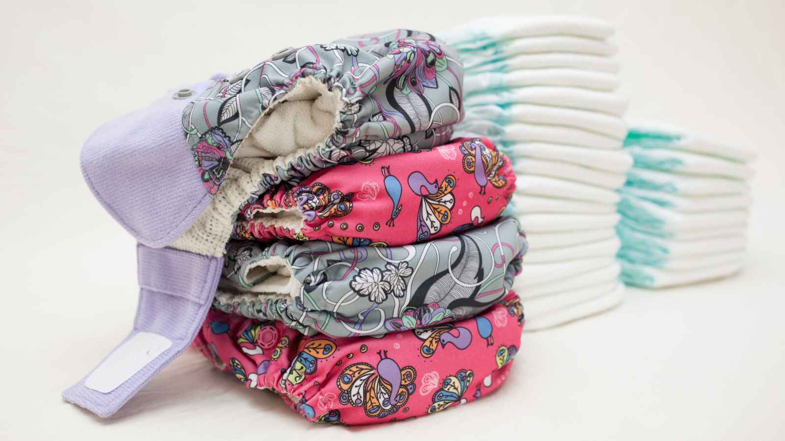 A stack of patterned cloth diapers is placed next to a pile of disposable diapers on a plain background.