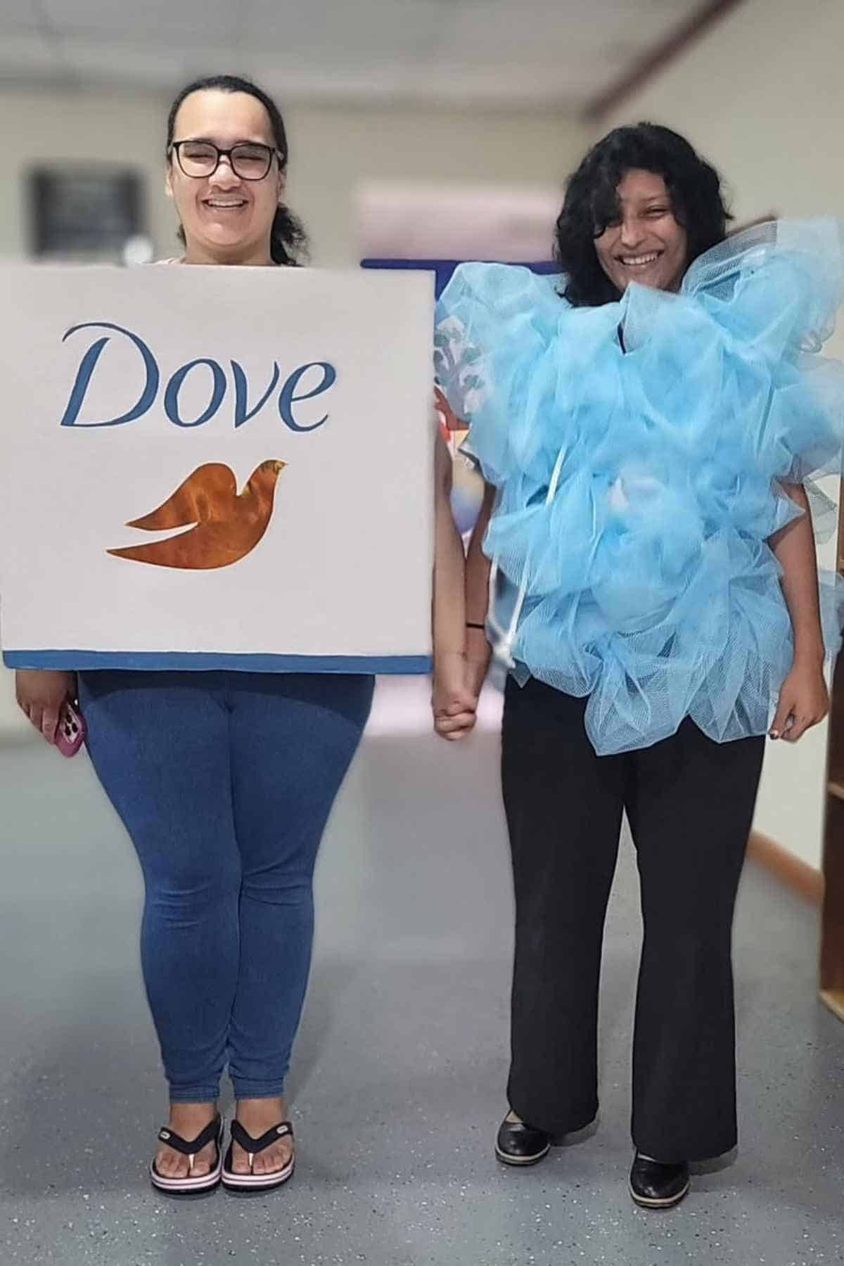 Two people stand indoors holding hands; one wears a box labeled "Dove" with a dove logo, and the other wears a blue costume resembling a shower puff.