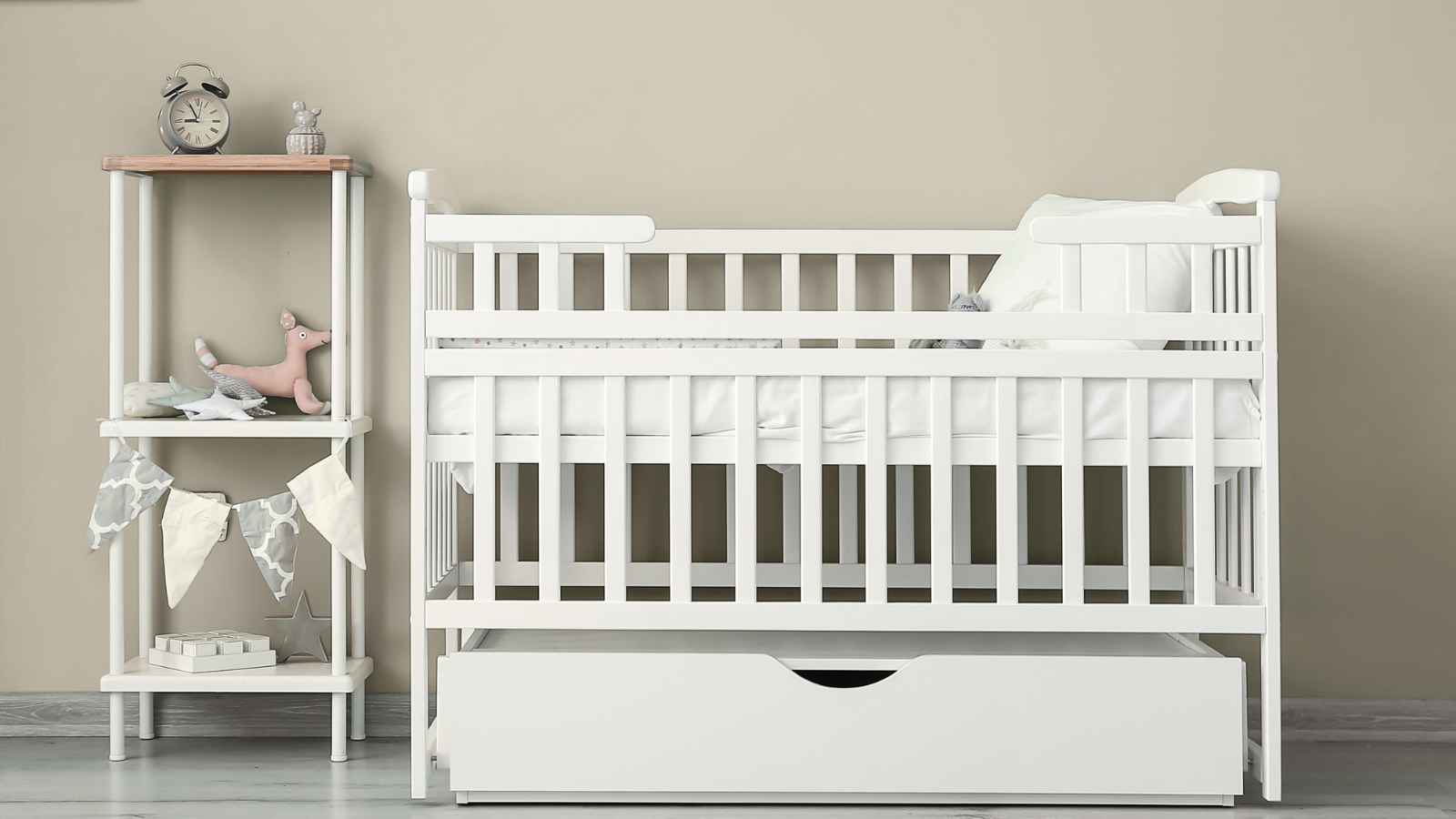 A white baby crib with a drawer underneath is placed next to a small shelf holding toys, books, and a clock against a beige wall.