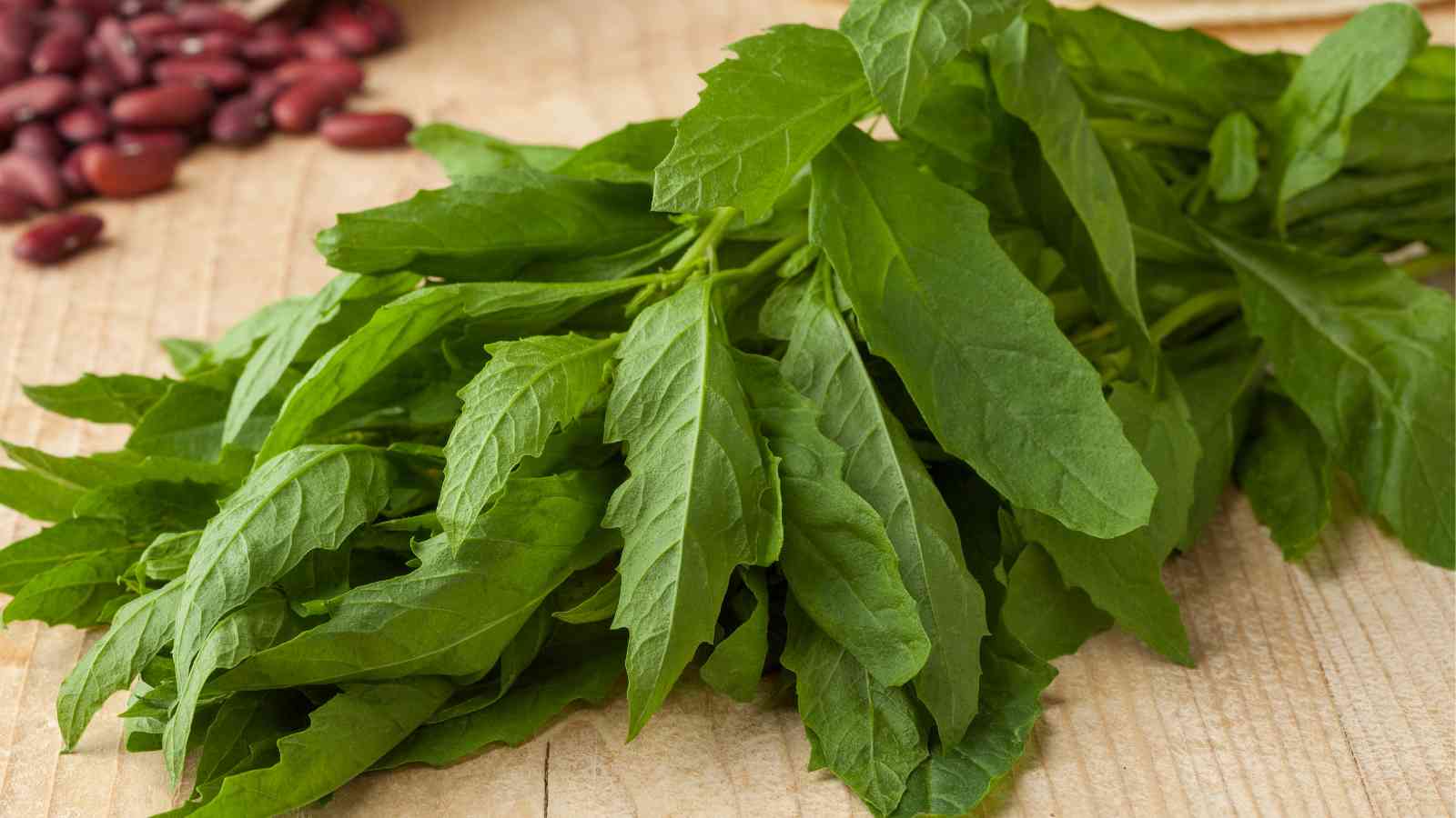 A bunch of fresh green epazote leaves placed on a wooden surface, with some red beans partially visible in the background.