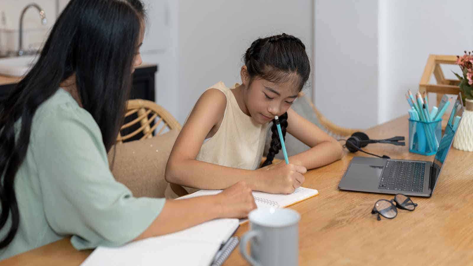 A woman and a young girl sit at a table with notebooks and a laptop; the girl is writing while the woman looks on.