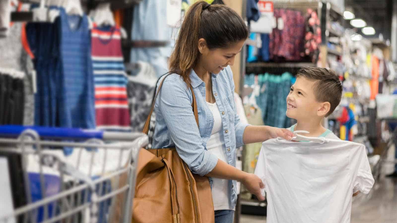 A woman and a boy stand in a clothing store aisle; the boy holds up a white T-shirt while the woman looks at him and smiles.