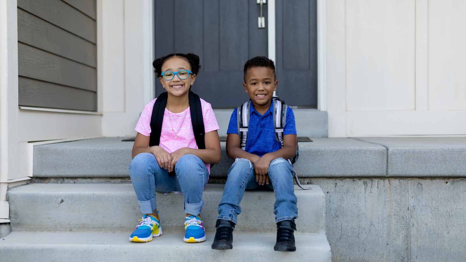 Two children with backpacks sit side by side on concrete steps in front of a house, smiling at the camera.