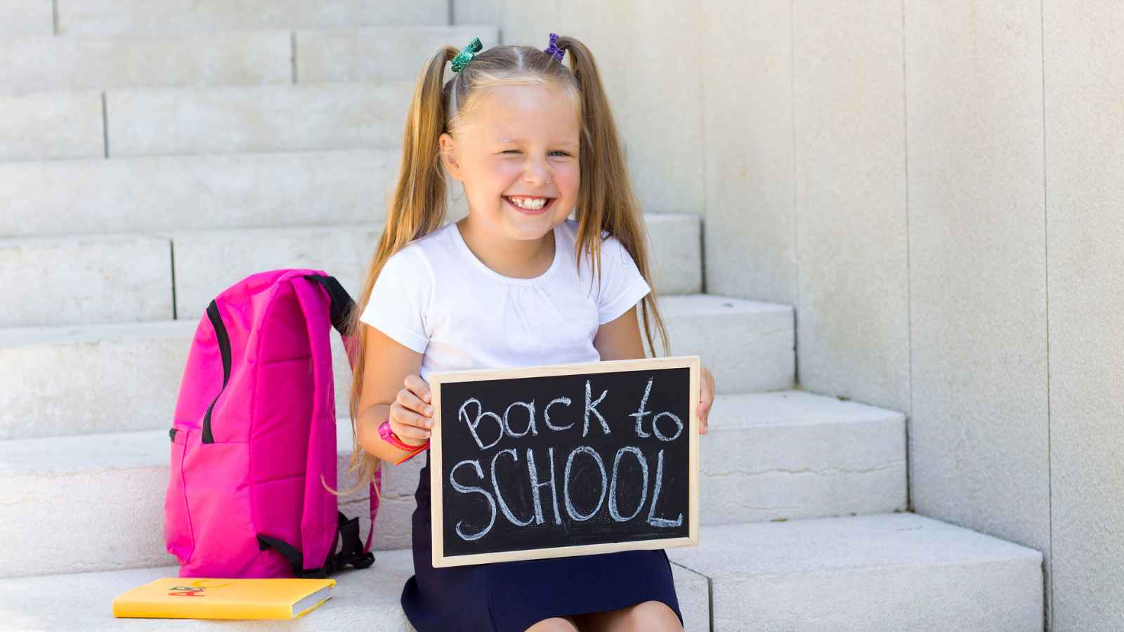 Young girl sitting on steps with a pink backpack and a notebook, smiling and holding a small chalkboard sign that says "Back to School.