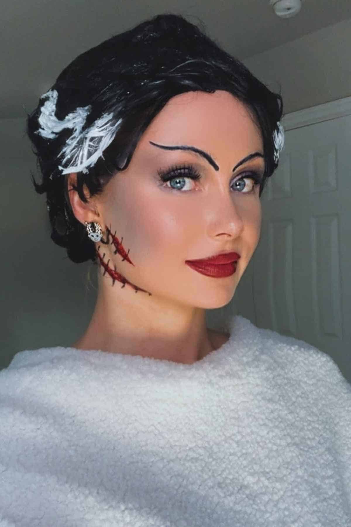 Woman wearing a white textured top and dramatic makeup, including black eyebrows, red lipstick, and fake stitches on her cheek, styled to resemble the Bride of Frankenstein.