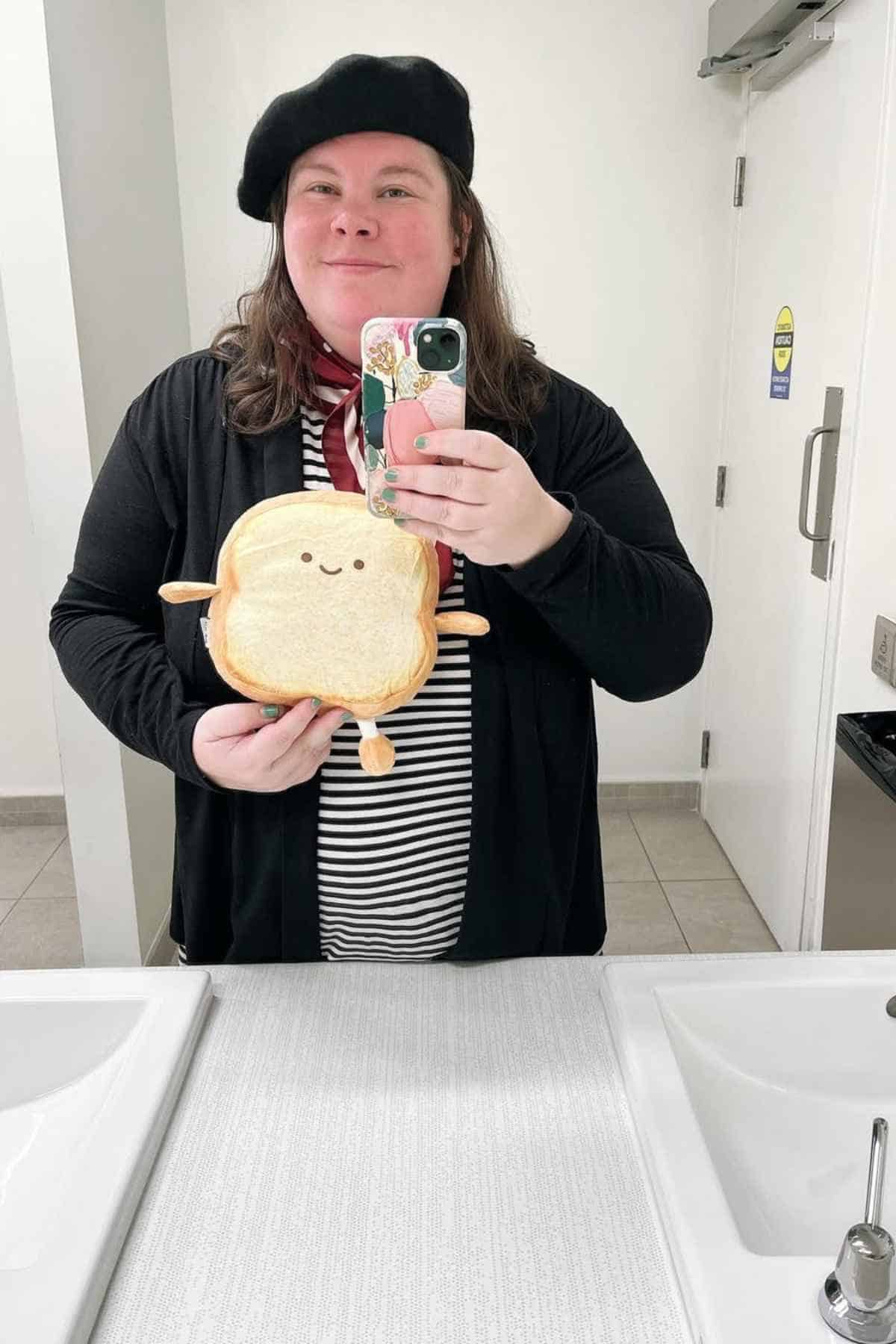 Person wearing a black beret and striped shirt takes a mirror selfie in a restroom, holding a plush toy shaped like a slice of bread with a smiling face.