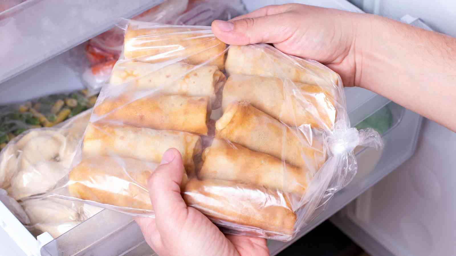 A person holds a plastic bag containing frozen spring rolls in front of an open freezer drawer with other frozen foods visible inside.
