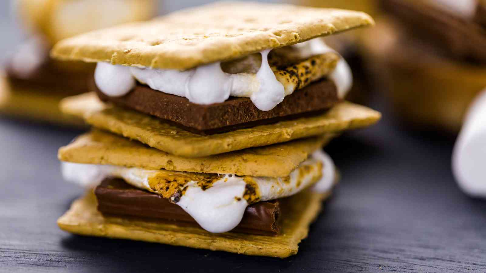 Two stacked s'mores made with graham crackers, toasted marshmallows, and chocolate pieces on a dark surface.