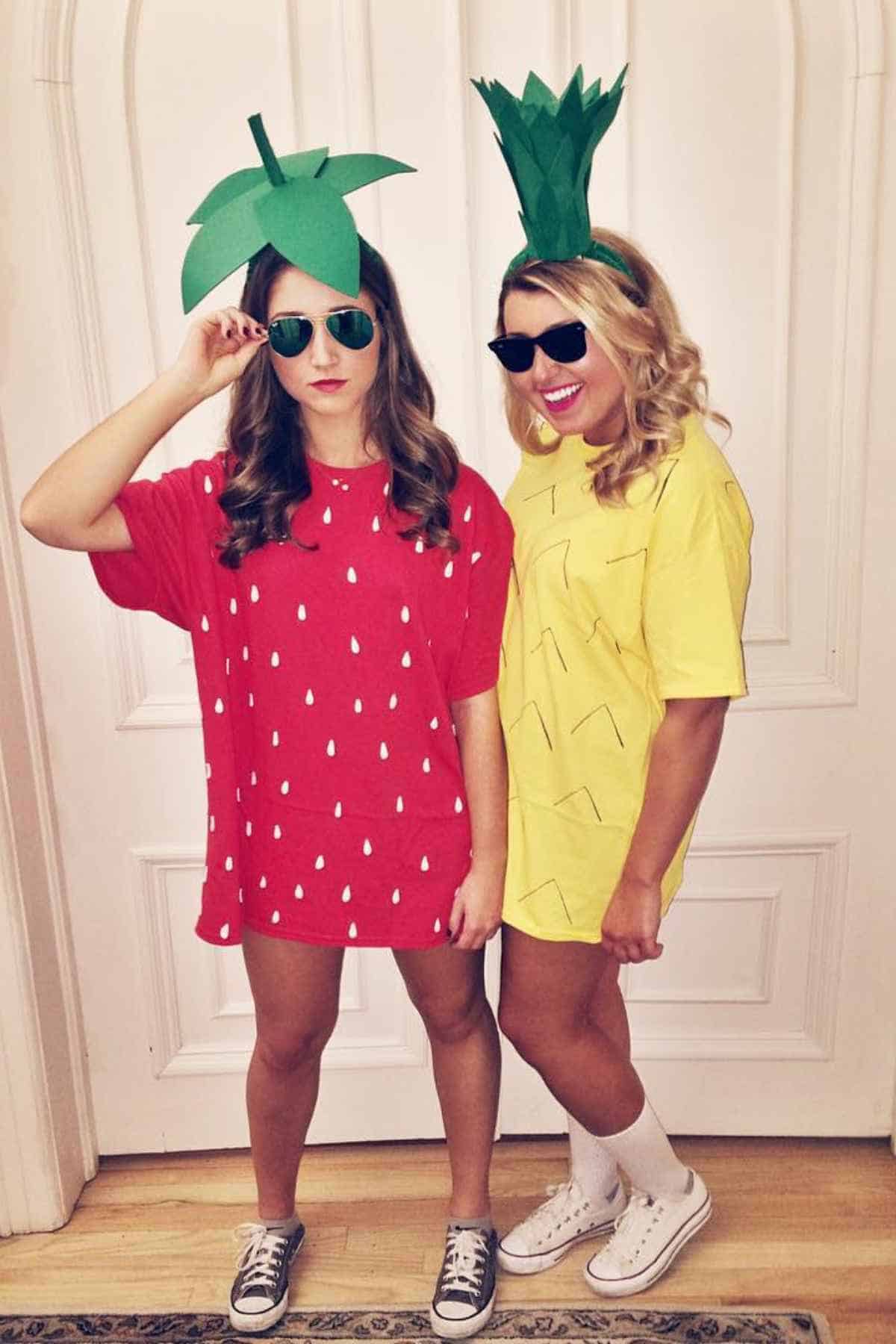 Two women wear fruit-themed costumes; one is dressed as a strawberry in red with a green leaf hat, and the other as a pineapple in yellow with a green crown hat. Both wear sunglasses.