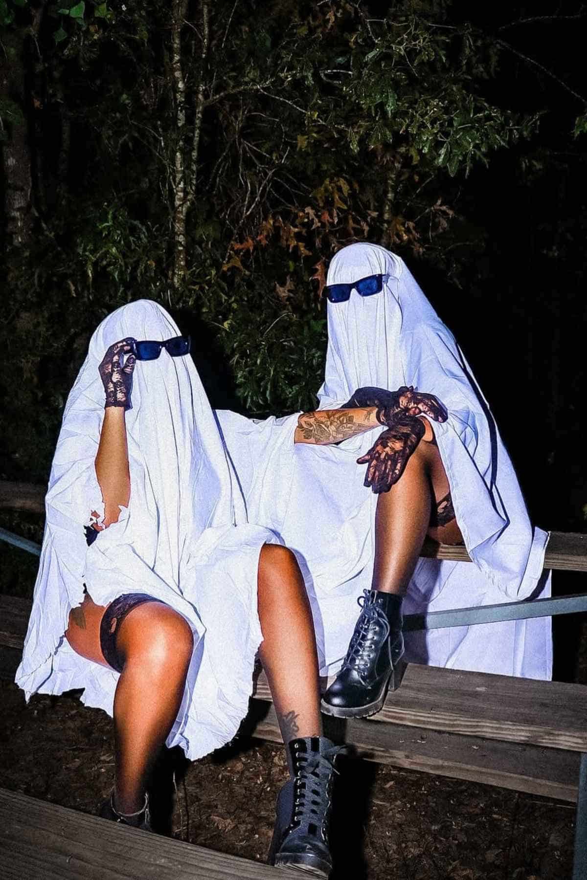 Two people dressed as ghosts with white sheets, sunglasses, and boots sit on a wooden bench outdoors at night.