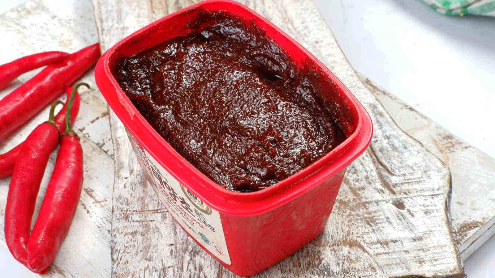 A red plastic container filled with gochujang sits on a wooden surface, with several whole red chili peppers nearby.