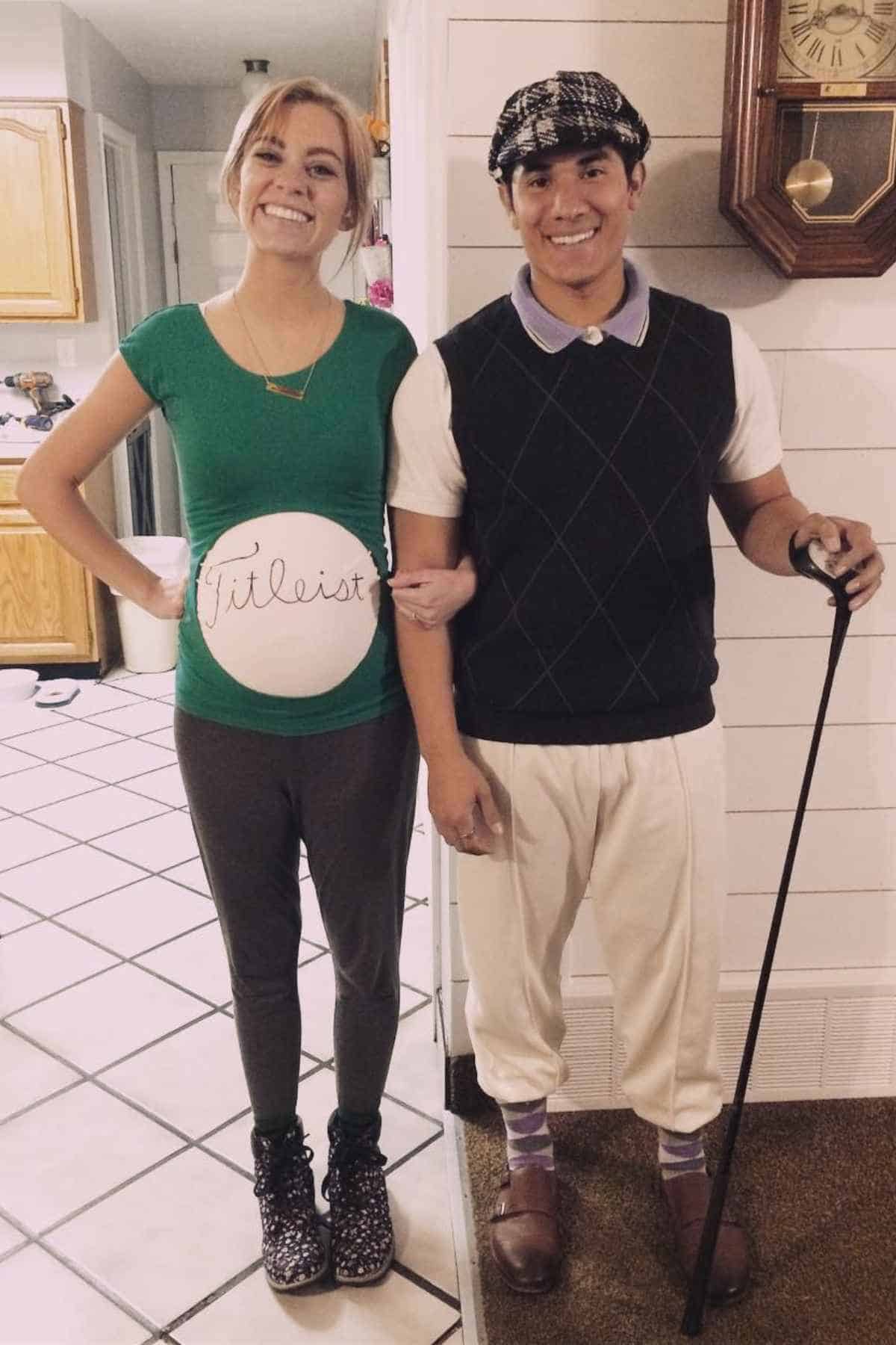 A smiling woman with a painted baby bump resembling a Titleist golf ball stands next to a man dressed in vintage golf attire, holding a golf club.