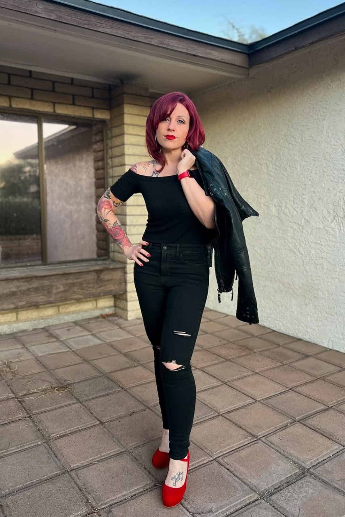 Person with red hair and tattoos poses on a patio in black off-shoulder top, ripped black jeans, red shoes, and a jacket draped over one shoulder.
