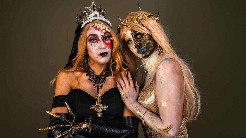 Two women in elaborate Halloween costumes and makeup pose against a plain background; one wears a black dress and crown, the other a gold outfit with a skeletal face design and crown.