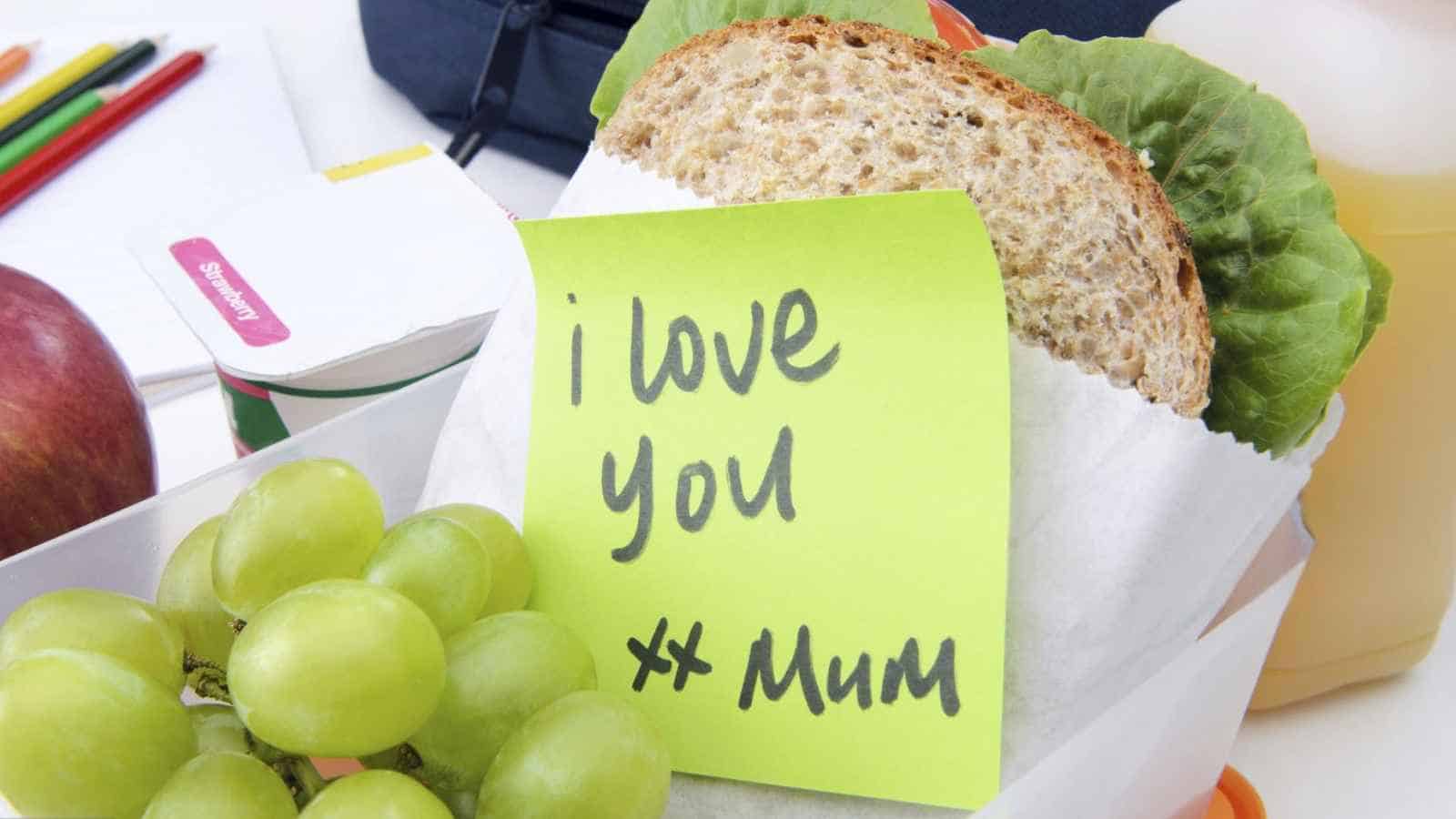 A sandwich in paper with a note reading "I love you xx Mum," next to green grapes and an apple in a lunchbox.