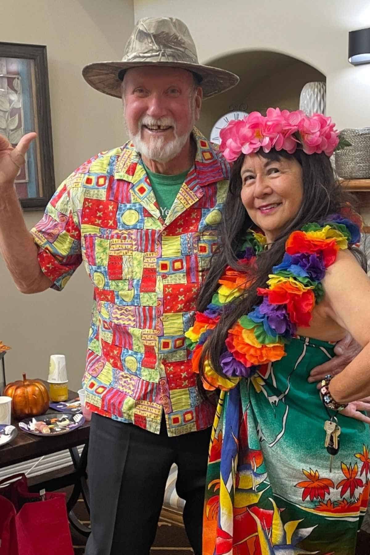 Two people dressed in colorful Hawaiian-themed outfits pose and smile indoors, with leis and floral accessories.