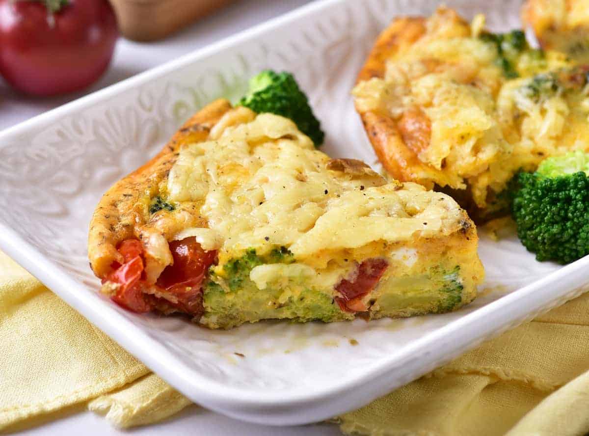 A slice of vegetable frittata with visible broccoli and tomatoes sits on a white plate.
