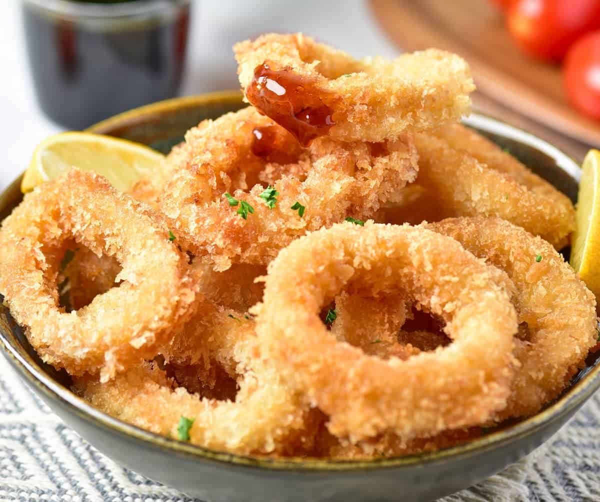 Crispy calamari fries served in a bowl with sliced lemons and a touch of sauce on one piece of calamari, plated on a mat.