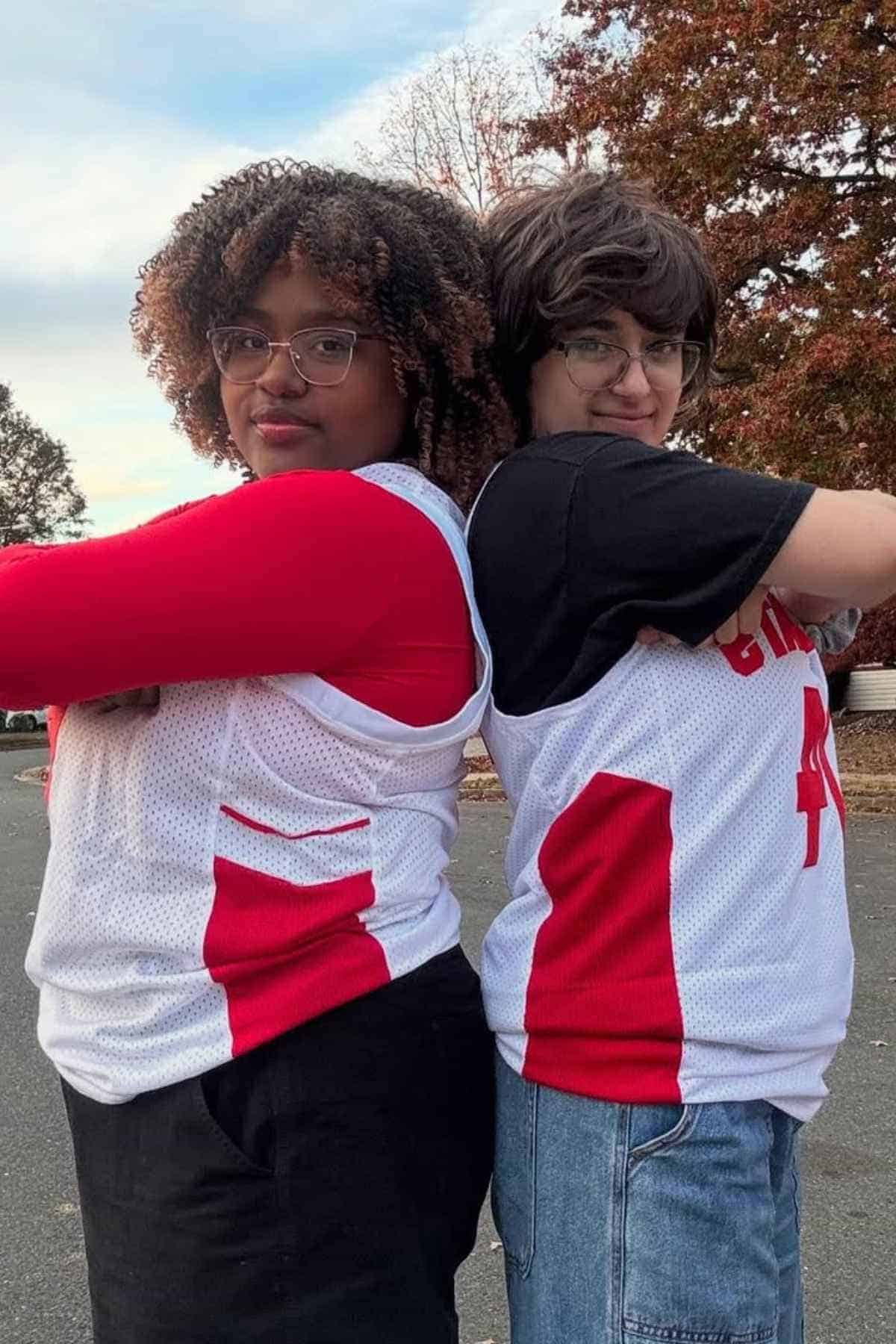 Two people stand back-to-back outdoors wearing white and red basketball jerseys, one in a red long-sleeve shirt and black pants, the other in a black T-shirt and denim pants.