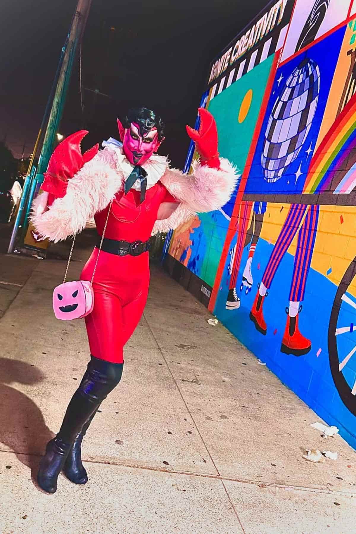 Person in a red and pink costume with a furry shawl poses on a sidewalk at night in front of a colorful mural featuring a disco ball, rainbow, and geometric designs, imitating Him in Power Puff Girls.