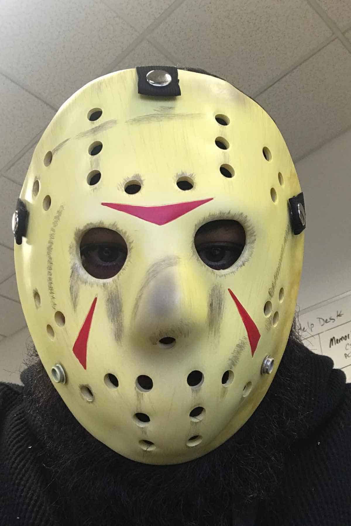 Person wearing a yellow hockey mask with red markings, similar to the one worn by the character Jason Voorhees, indoors under fluorescent lighting.