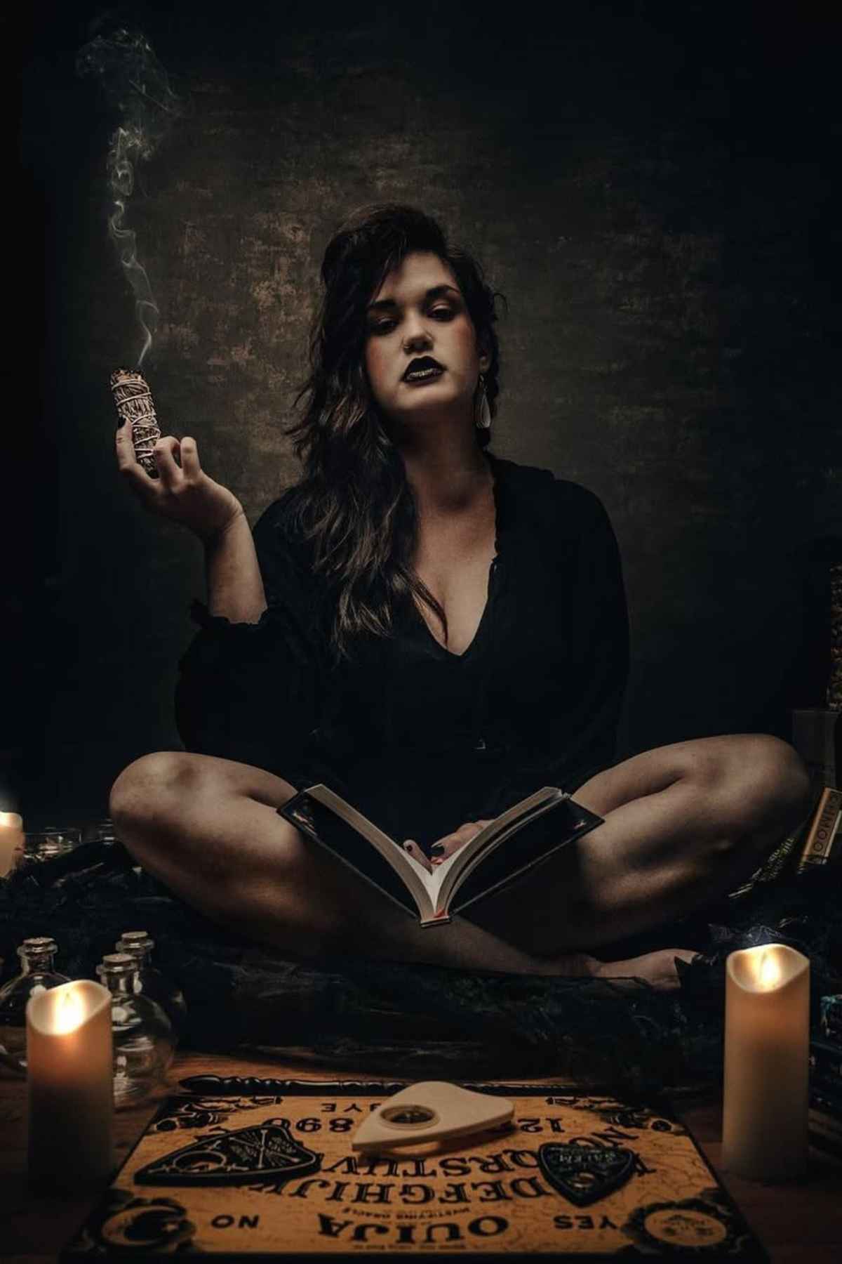 A woman sits cross-legged in dim lighting, holding a smoldering bundle, with an open book, candles, and a Ouija board arranged in front of her.