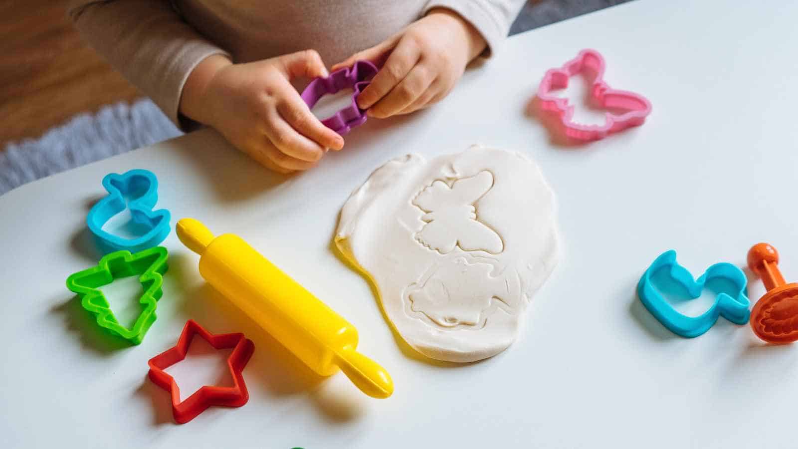 A child uses cookie cutters and a rolling pin to shape dough on a table, with various colorful cutters and shapes scattered around.